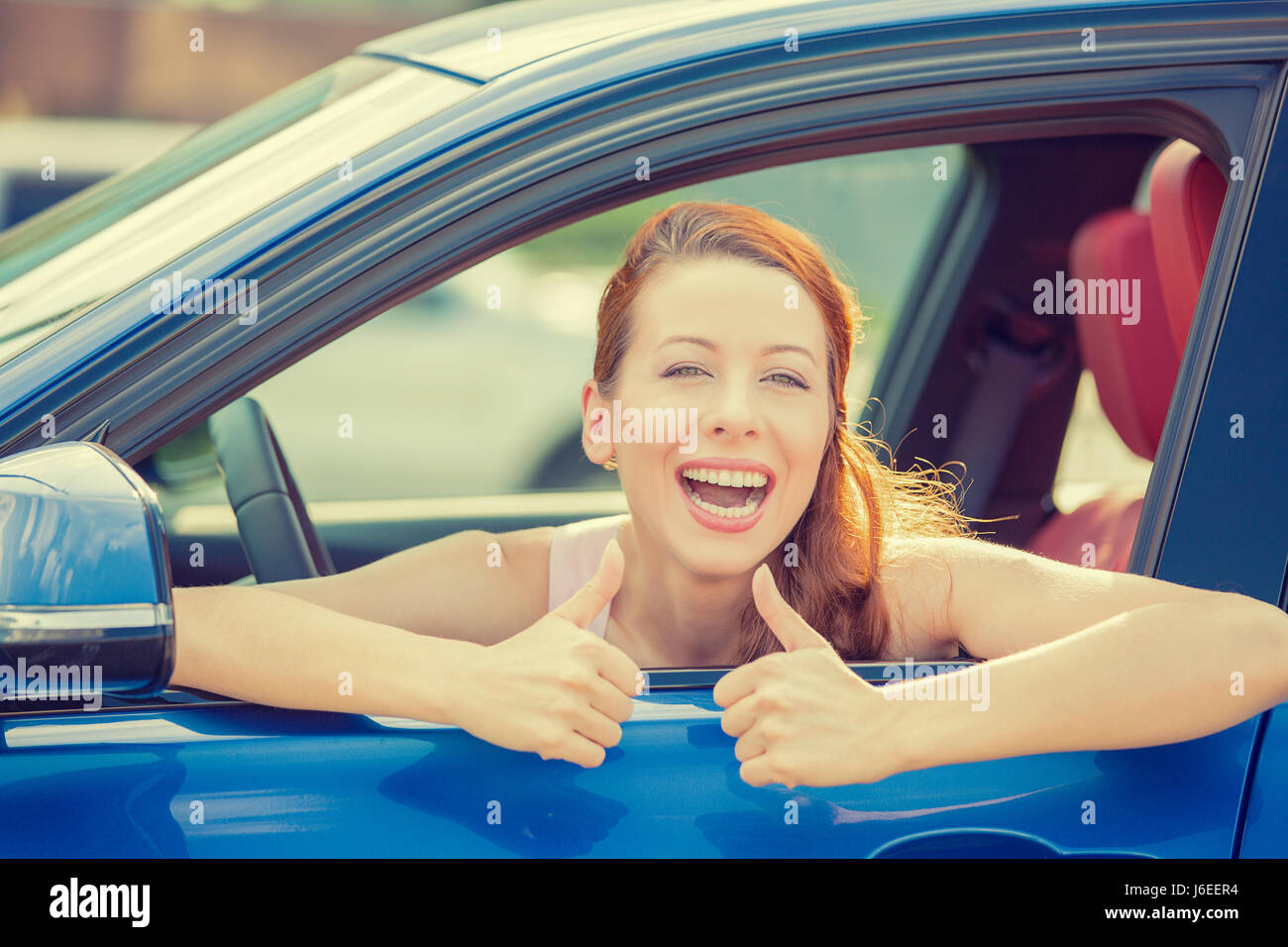 Side door view woman driver happy smiling showing thumbs up sitting inside new blue car  outside on parking lot background. Beautiful young woman happ Stock Photo