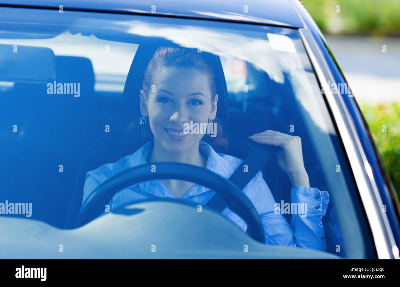 Closeup portrait through windshield window, smiling young attractive woman pulling on seatbelt inside black car. Driving safety, buckle up to prevent  Stock Photo