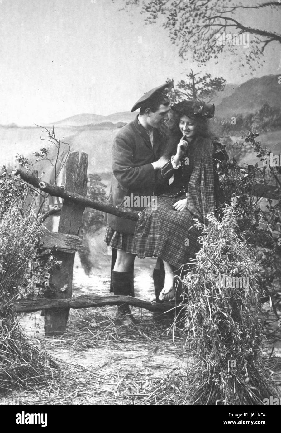 Scottish man in full tartan outfit, flirts with a tartan-dressed woman who sits on a wood fence in the countryside. He is gently embracing her, she is shyly smiling. 1911.   To see all my Alamy  vintage images - in Search, key these 2 words:  Prestor  vintage Stock Photo