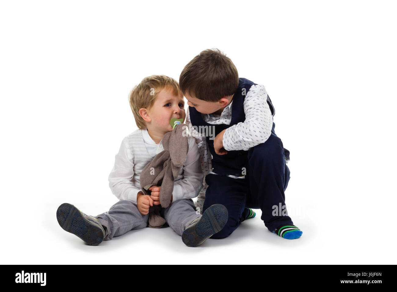 2 Little brothers wearing festive clothing sitting together on the floor, toddler has plush animal (rabbit) and pacifier. Isolated on white background Stock Photo