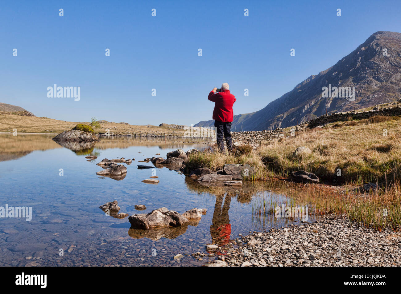 Senior man in red fleece jacket taking photos on his cellphone, at Llyn Idwal, Snowdonia, North Wales, UK, on a beautiful sunny spring day with clear  Stock Photo