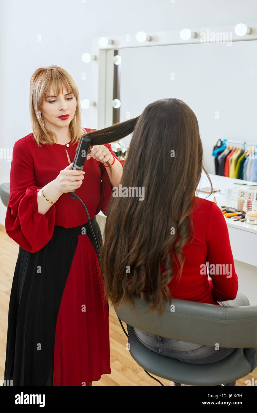 professional stylist using curling iron and doing curls women's hair. Hair curls in beauty salon. Backstage photo stylist working in studio dressing r Stock Photo