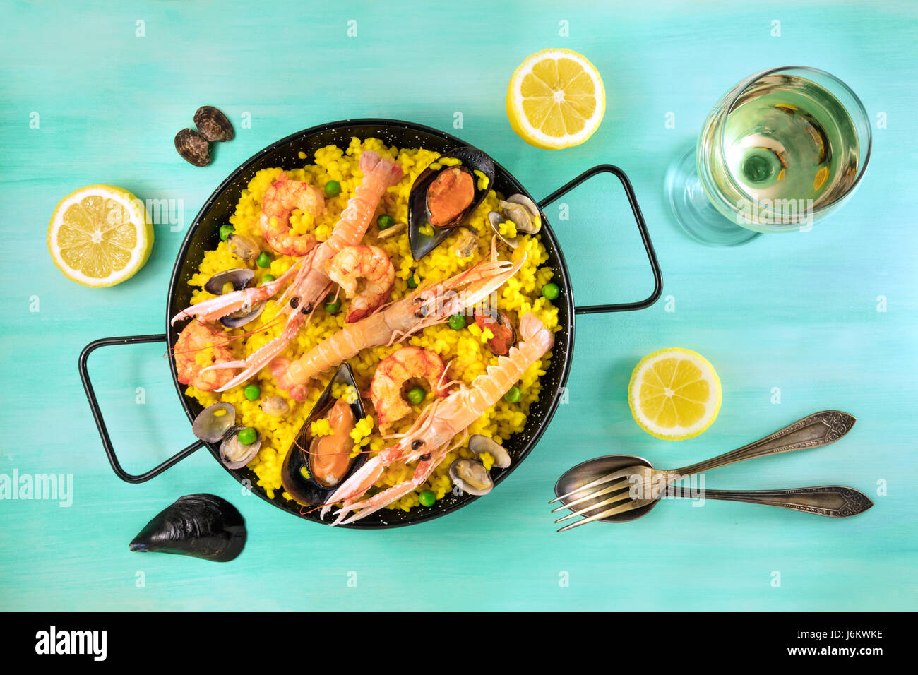 Photo of Spanish seafood paella in typical paellera, with slices of lemons, mussels and clams shells, glass of white wine, fork and spoon, and a place Stock Photo