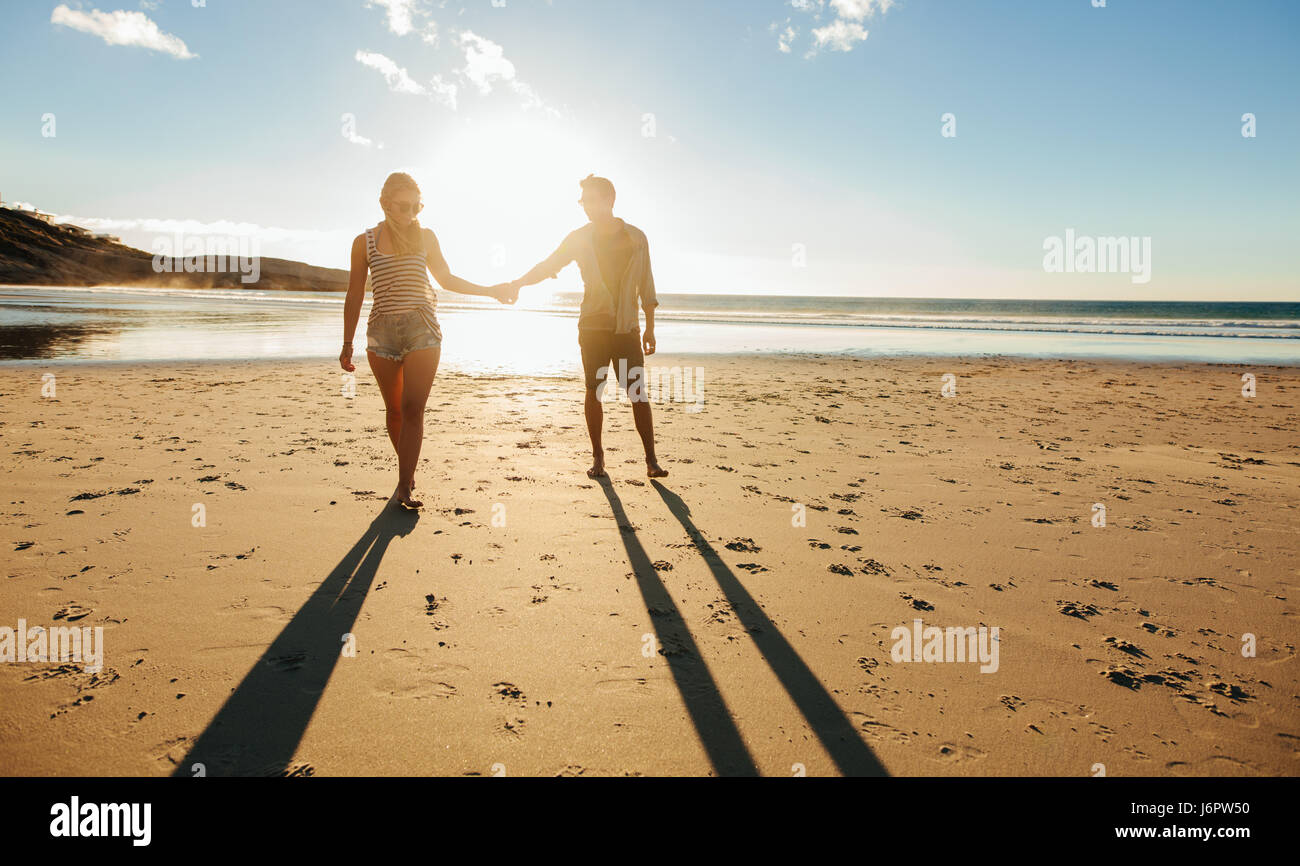 Outdoor shot of romantic young couple walking along the sea shore holding hands. Young man and woman walking on the beach together at sunset. Stock Photo