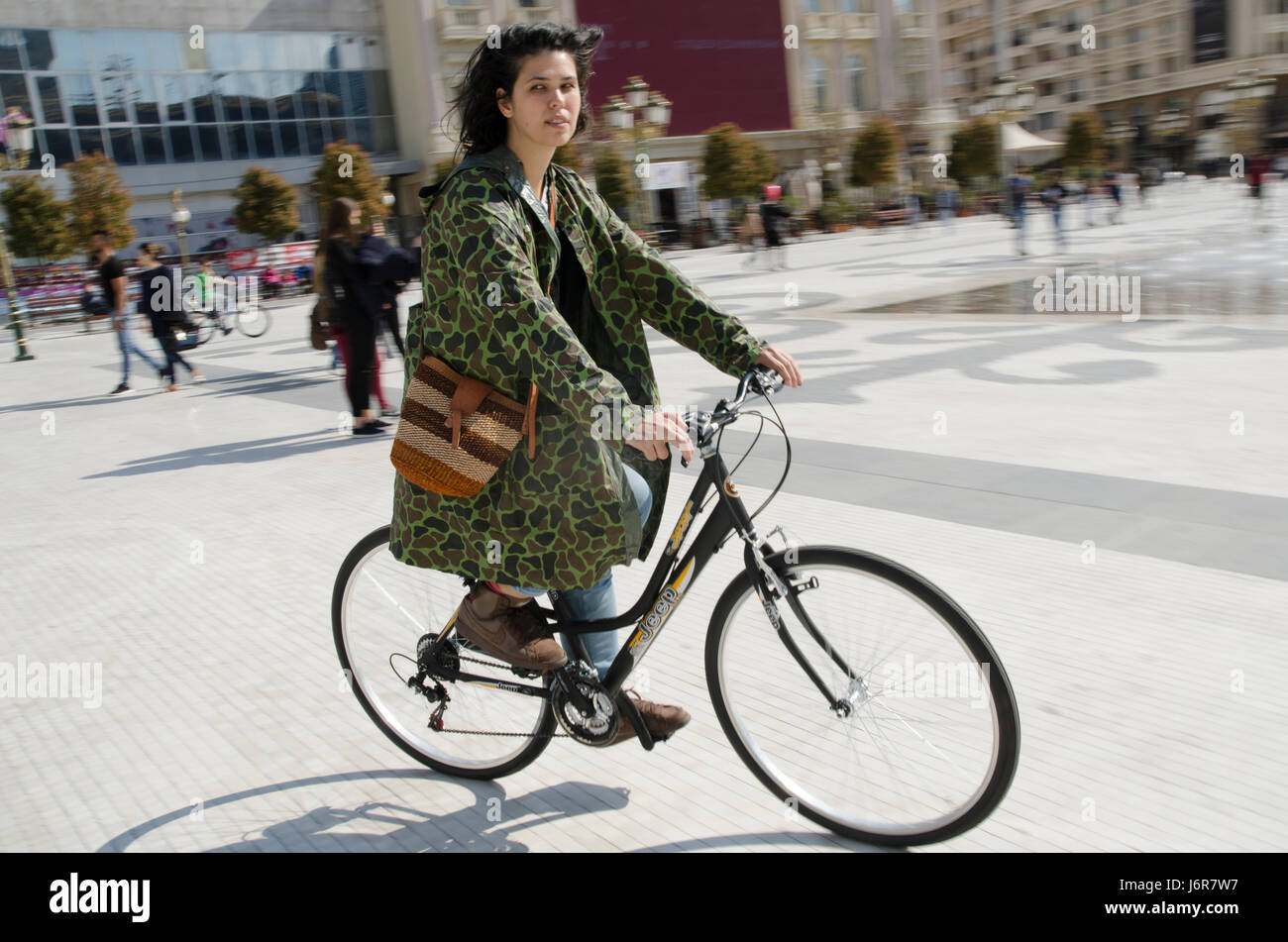 Woman with camouflage raincoat riding bicycle in Skopje square. Stock Photo