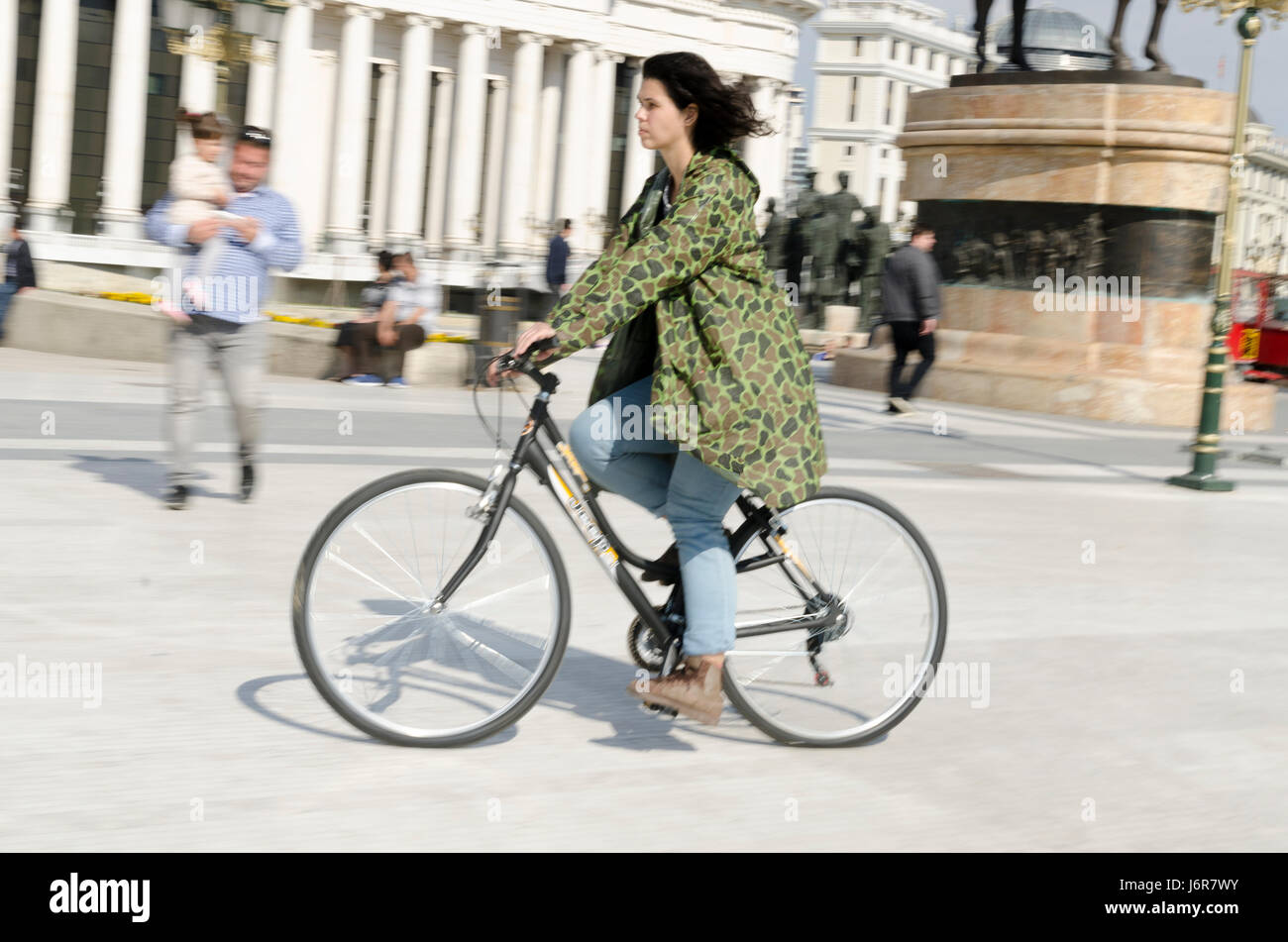 Woman with camouflage raincoat riding bicycle in Skopje square. Stock Photo