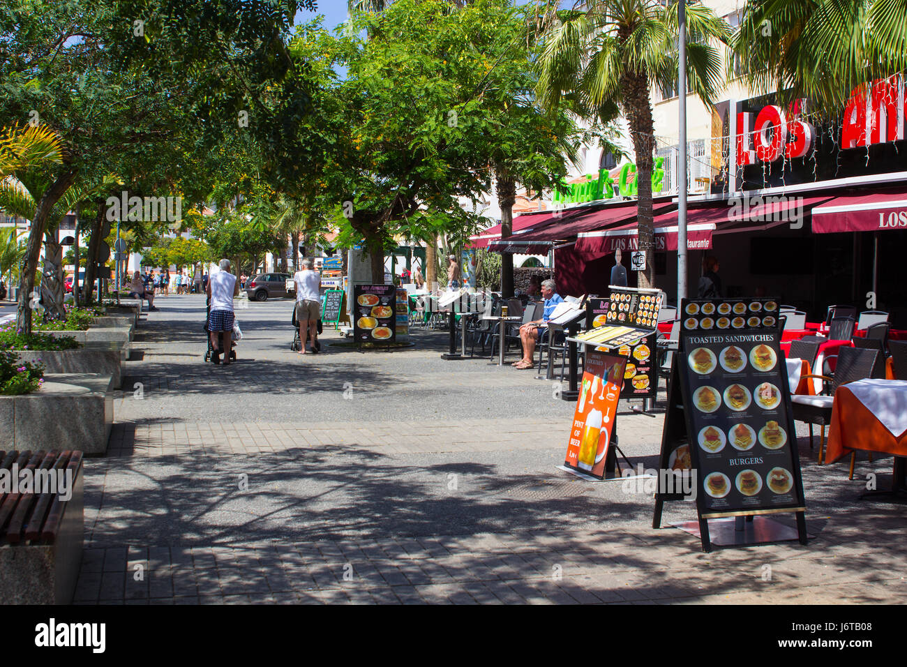 Tourists walking and dining on a tree lined avenue with shops and cafes in Playa de Las Americas on the island of Teneriffe Stock Photo