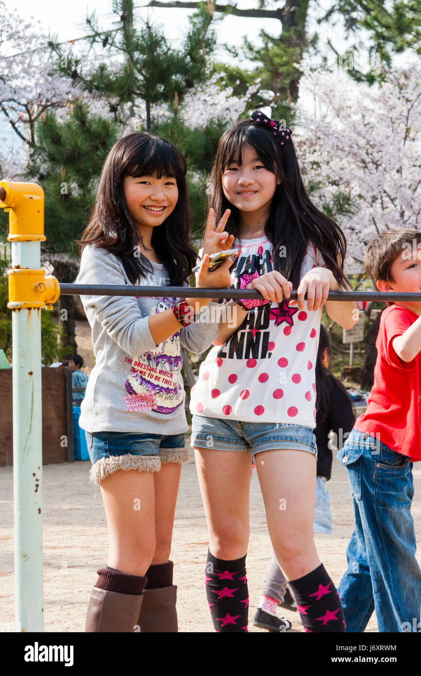 Two young teenage Japanese girls, 12-13 years old,  in t shirts and cut down jeans smiling and giving peace sign to viewer, cherry blossoms behind. Stock Photo