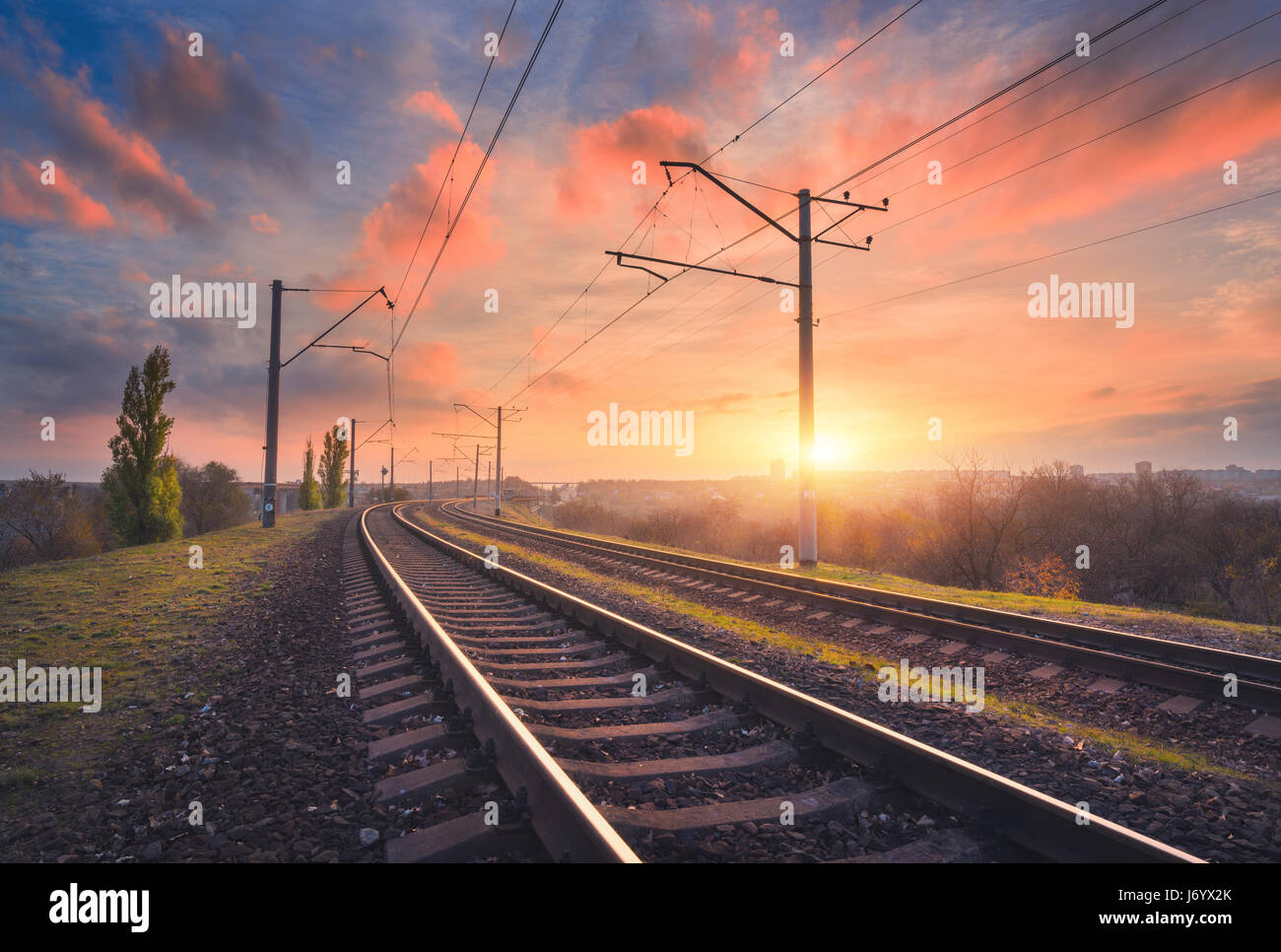 Railroad and beautiful sky at sunset. Industrial landscape with railway station, colorful blue sky with red clouds, trees and green grass, yellow sunl Stock Photo