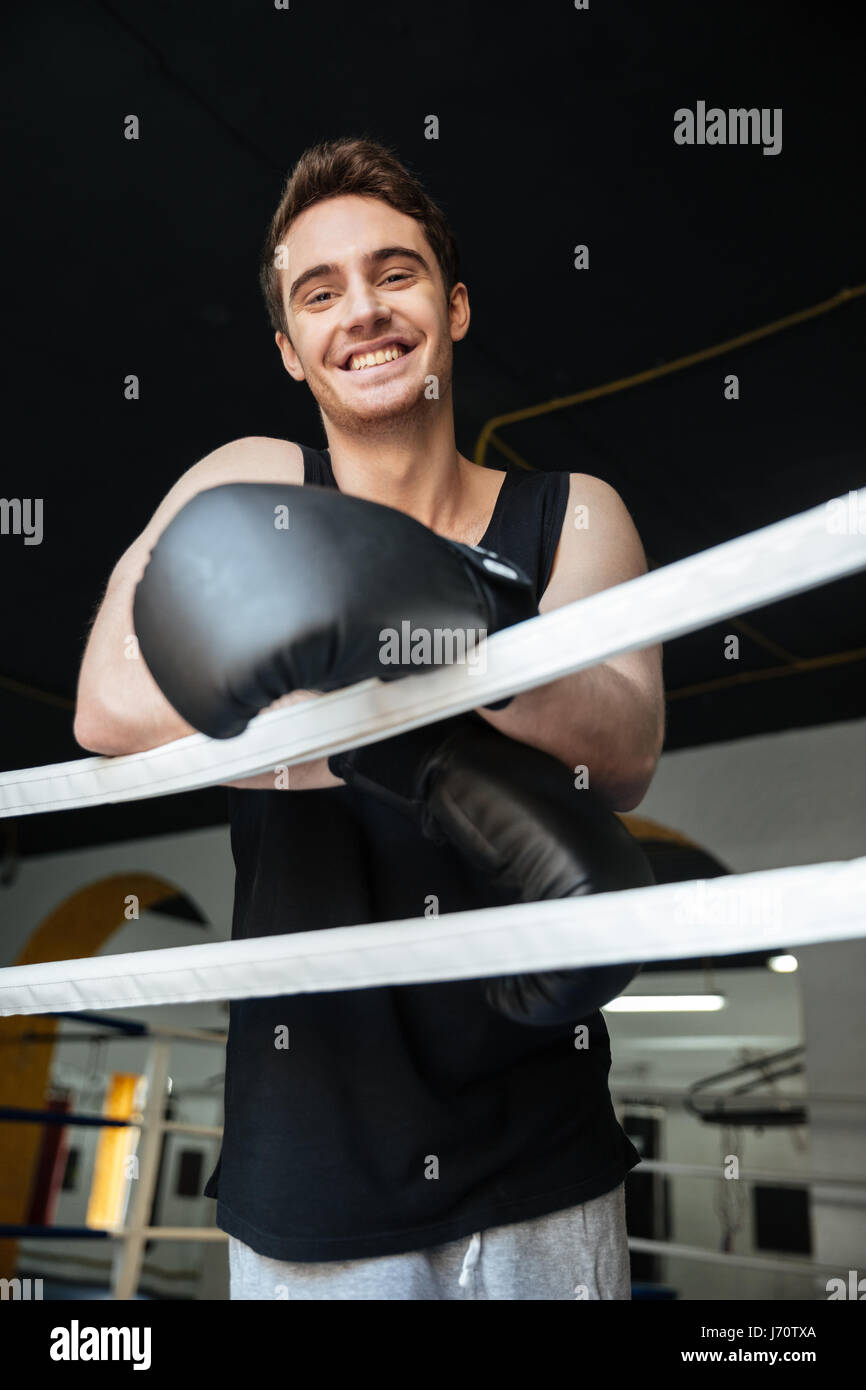 Cheerful boxer wearing black boxing gloves looking at camera in gym ...