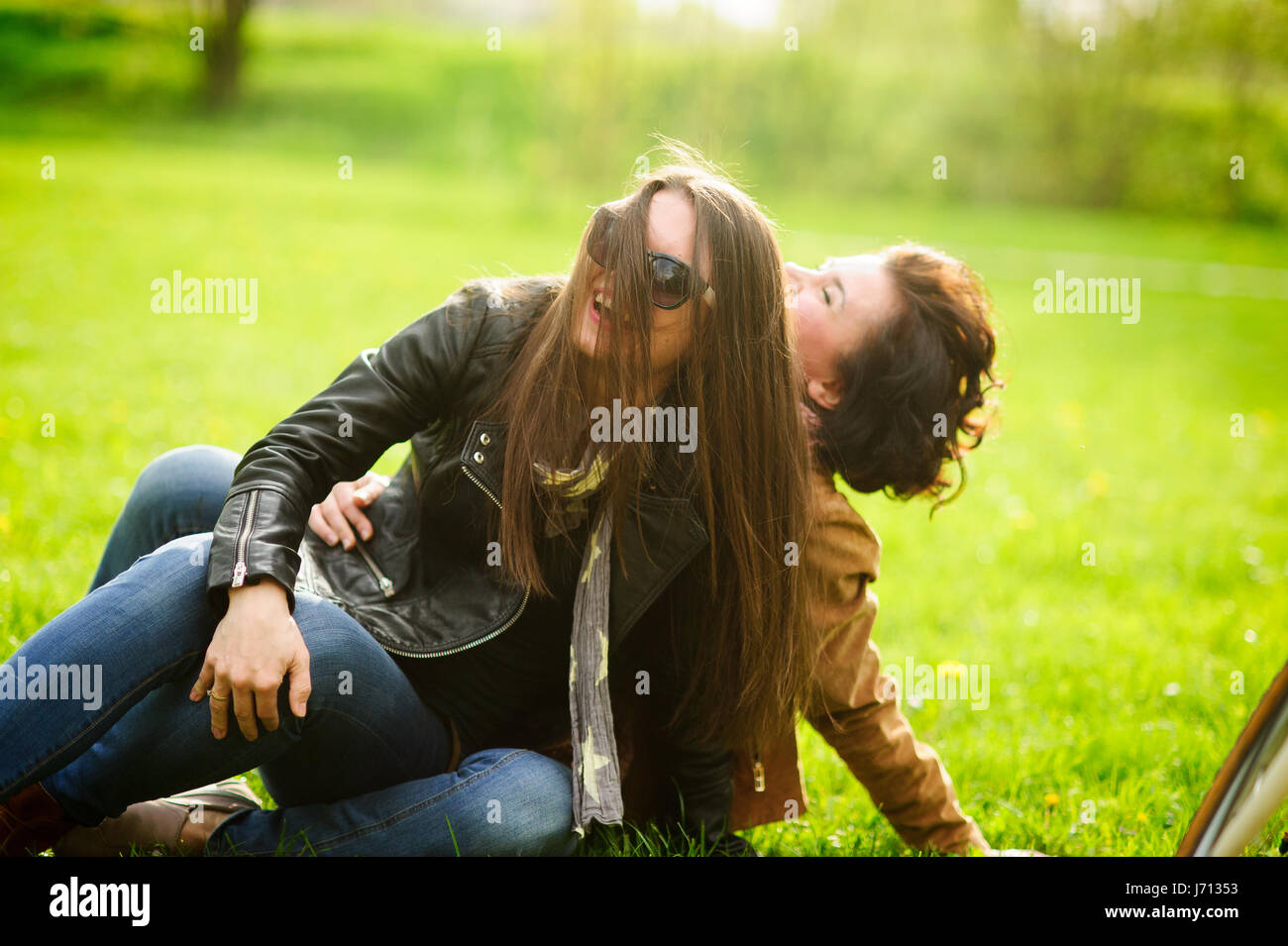 Two pretty young women cheerfully spend time in the spring park. Girlfriends frolicking while sitting on the green grass. Sunny warm day. Good mood. Stock Photo