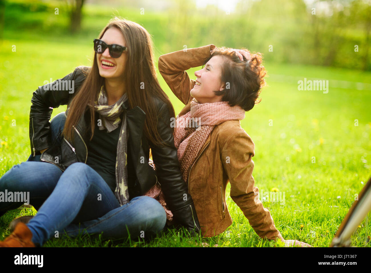 Two pretty young women cheerfully spend time in the spring park. Girlfriends frolicking while sitting on the green grass. Sunny warm day. Good mood. Stock Photo