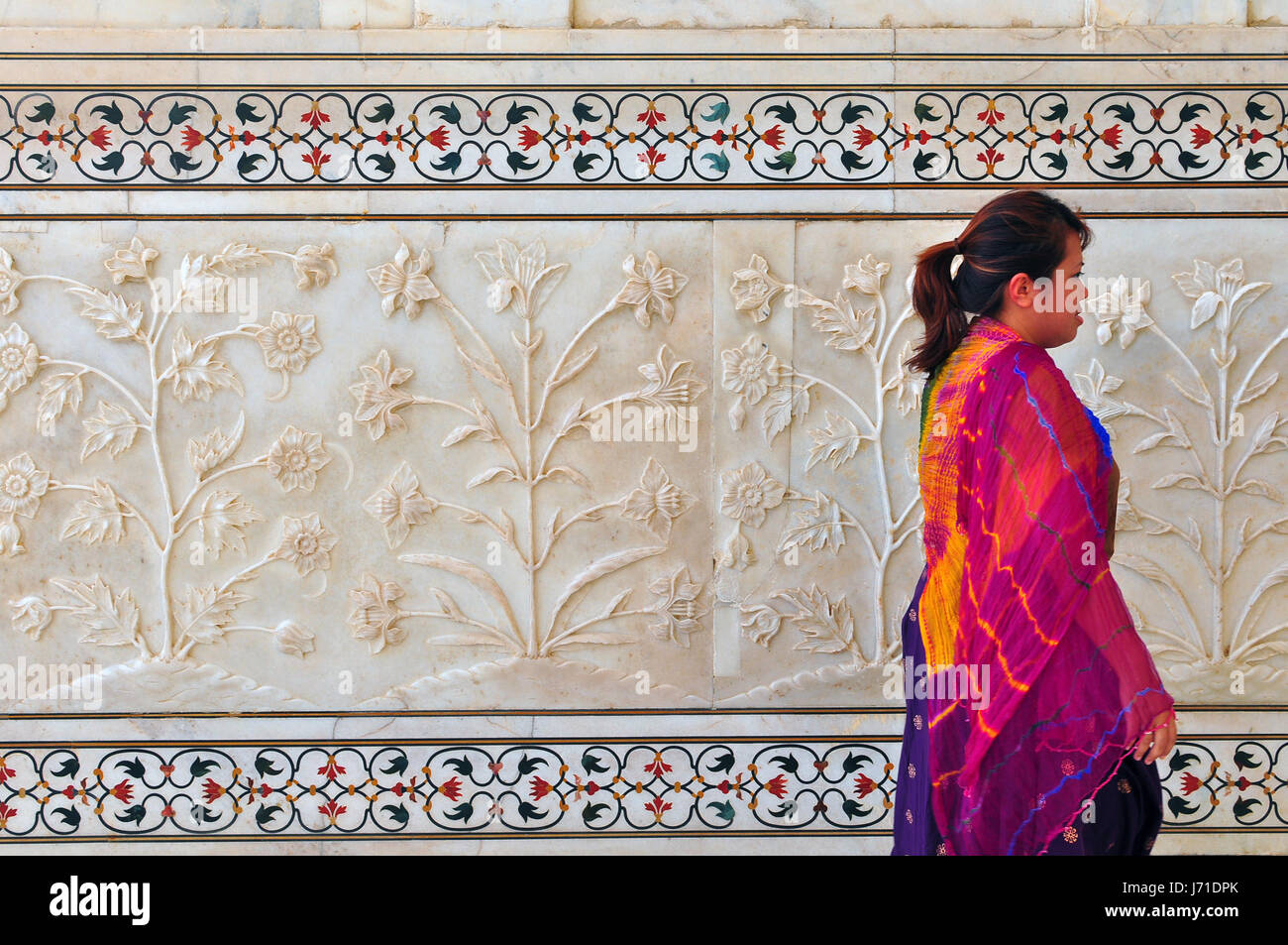 A girl with a colourful shawl walks past an intricately carved marble wall with inlaid stones. Stock Photo