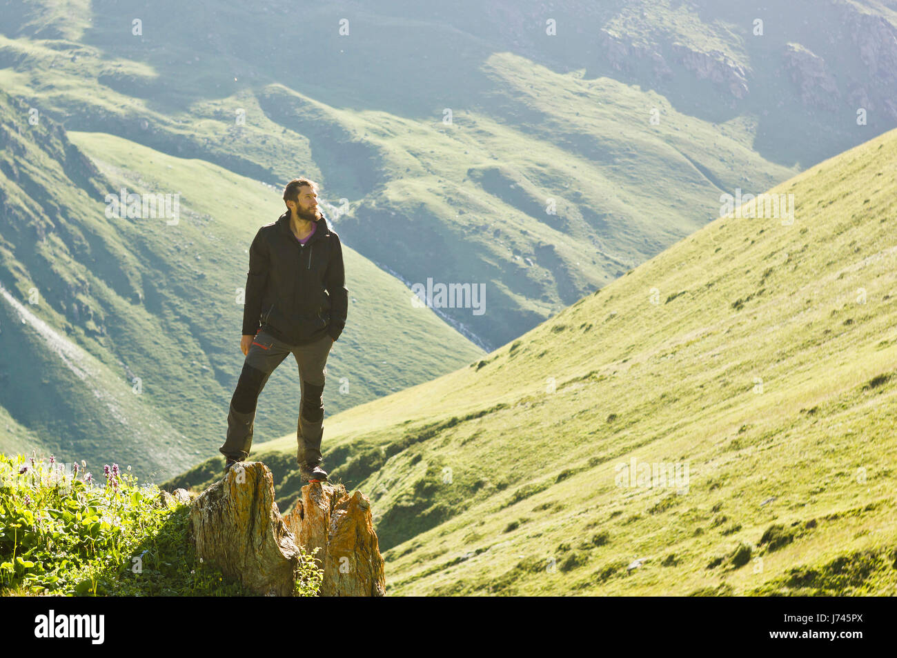 man in black jacket standing on stone with hands-up above mountais at sunset Stock Photo