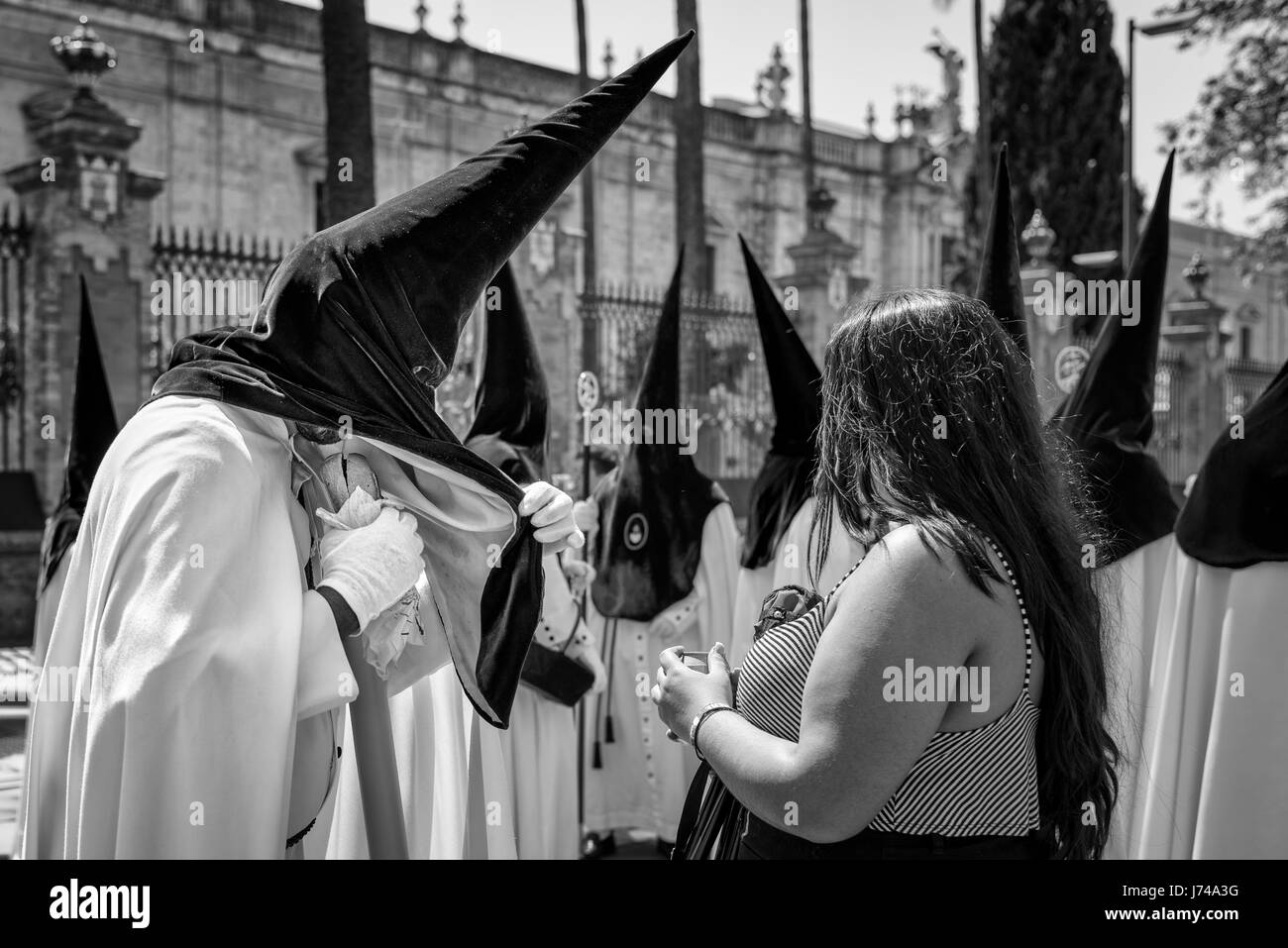 Woman feeding a Nazarene penitent during a break in the procession. Processions last for hours and the participants need to get drinks and food withou Stock Photo