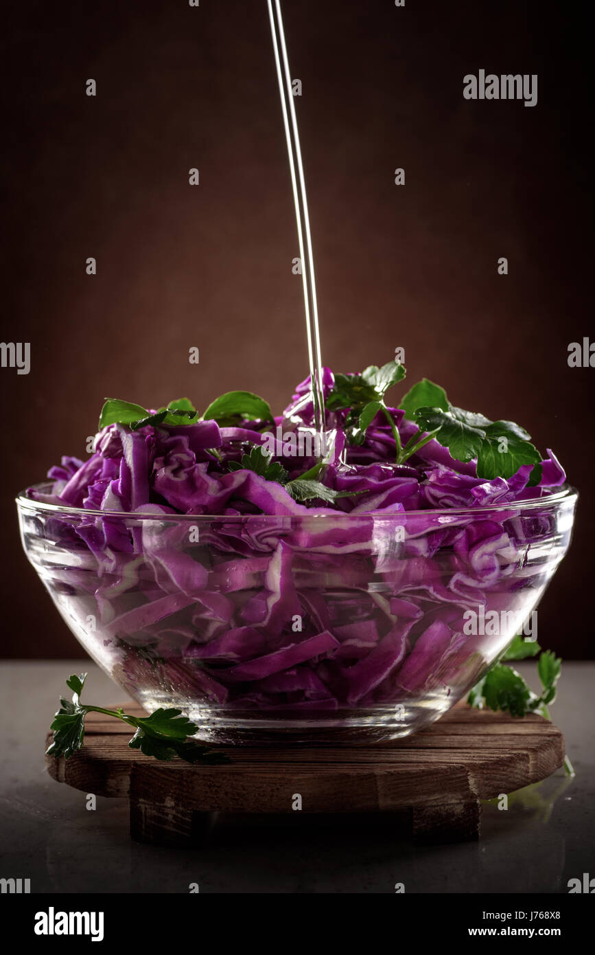 olive oil in a thin stream pour into Salad of red cabbage in glass transparent bowl on dark background Stock Photo
