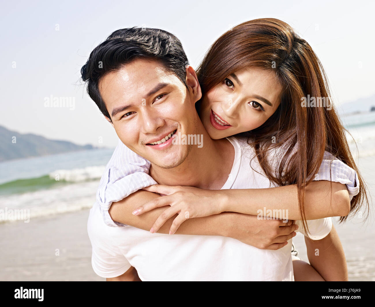 young asian man carrying girlfriend or wife on back on beach. Stock Photo