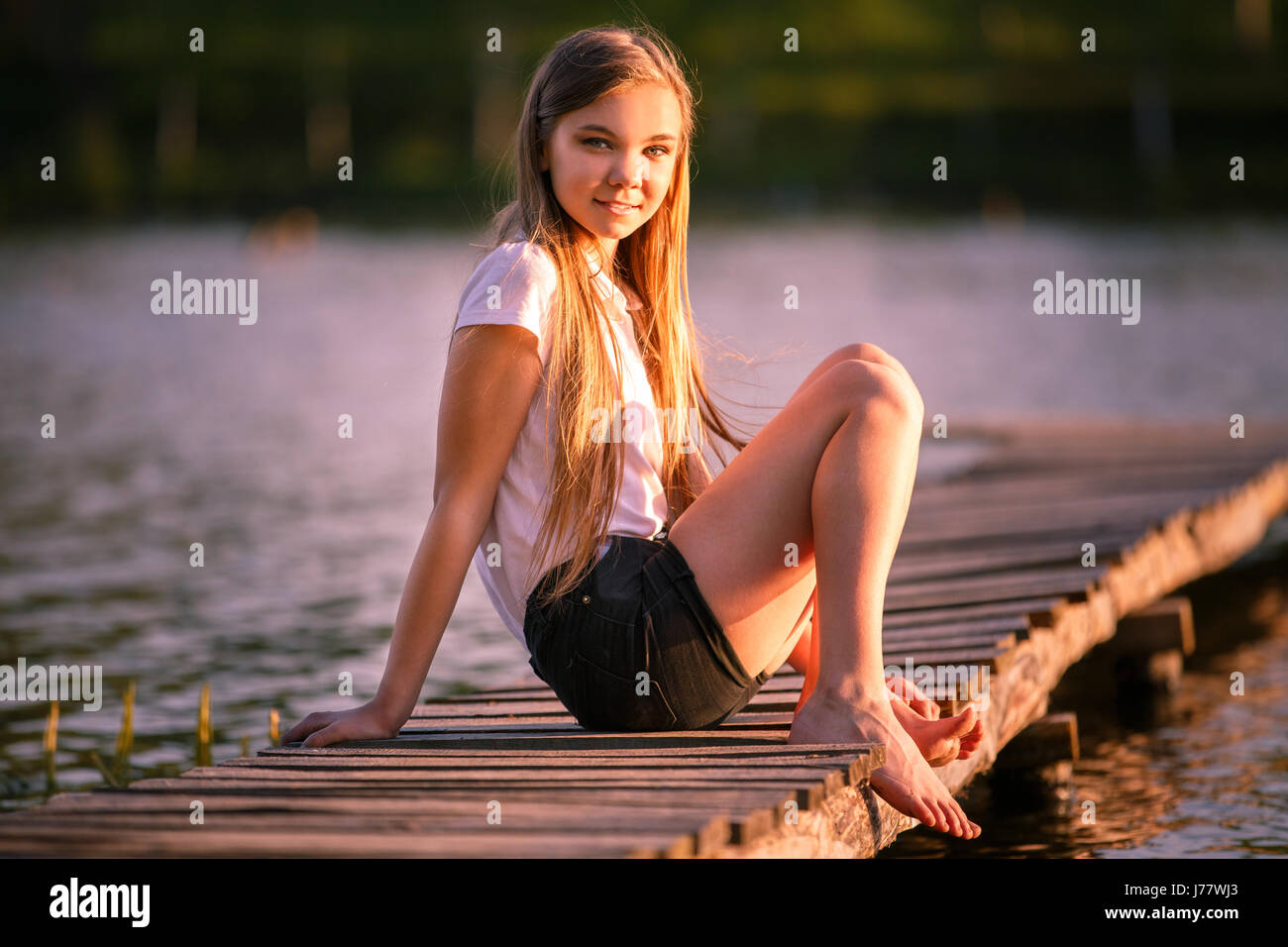 Young smiling teenage girl sitting on pier in sunset beams Stock Photo