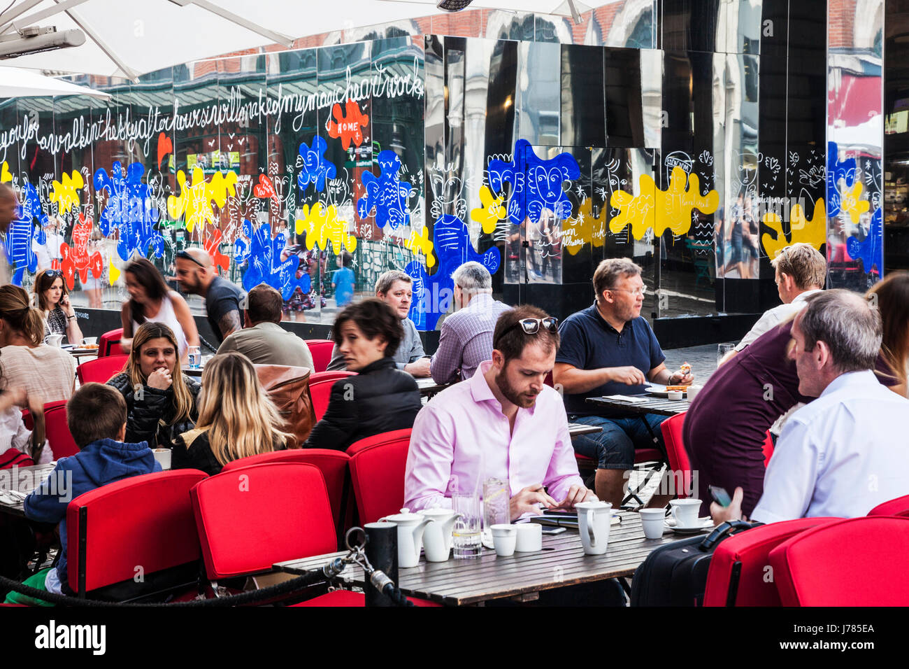 People sitting at an outdoor cafe in Covent Garden with the mirrors of Reflect London behind. Stock Photo
