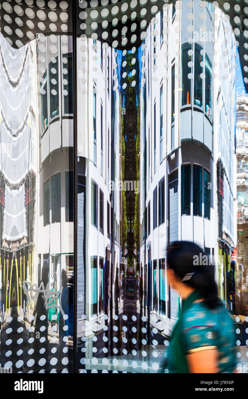 Asian woman walking past an office building looking at the distorted reflections in the windows. Stock Photo