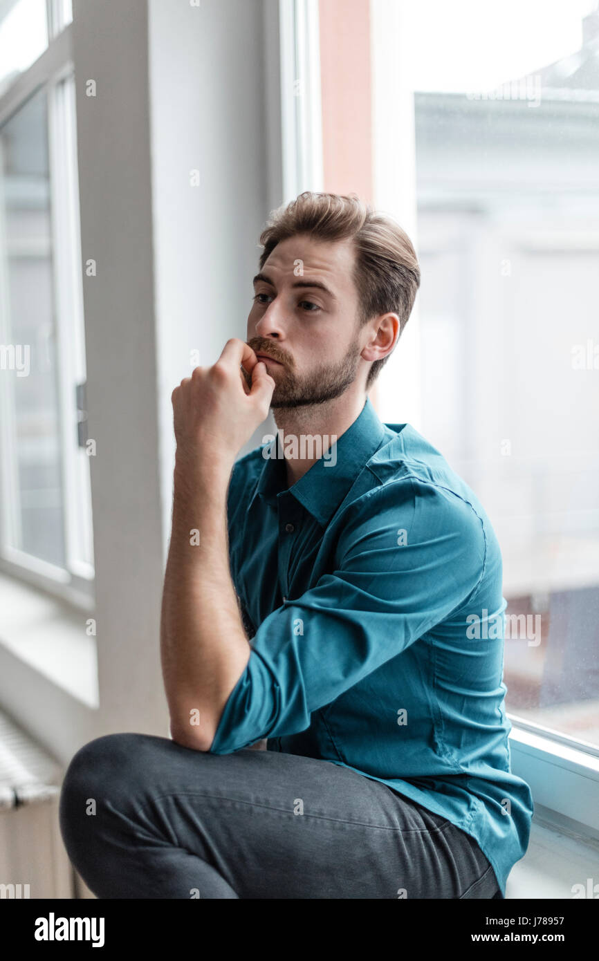 Portrait of pensive young man sitting on window sill Stock Photo - Alamy