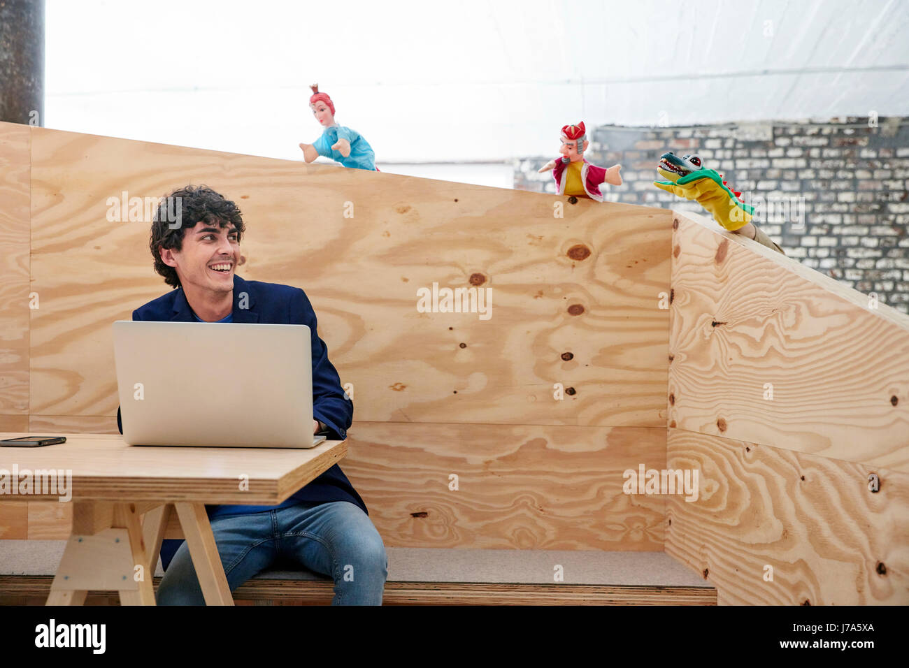 Young businessman working on laptop with hand puppets watching him from wooden wall Stock Photo