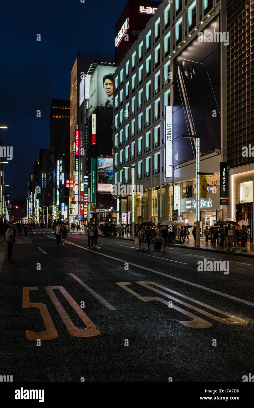 Nighttime photo of a busy downtown area showcasing night lights and pedestrians waiting or walking along the pavement or crossing the street. Stock Photo