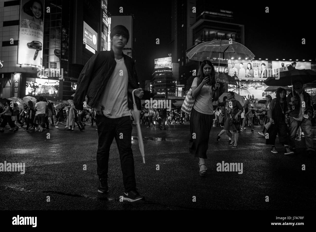 Black and white night photo of pedestrians on the busy Shibuya Crossing. A girl, texting and carrying an umbrella, walks slightly behind a young man. Stock Photo