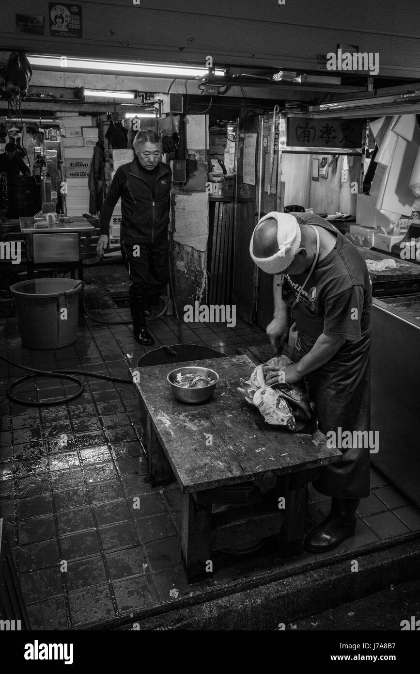 Black and white photo of two men working in their stall at the Tsukiji Fish Market. The fishmonger is emptying and cleaning a big fish head. Stock Photo