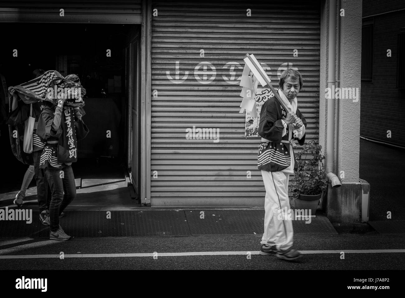 Two men walking down a street: the man in front is carrying a paper flag and is followed by another one wearing a dragon head mask.  Black and white. Stock Photo