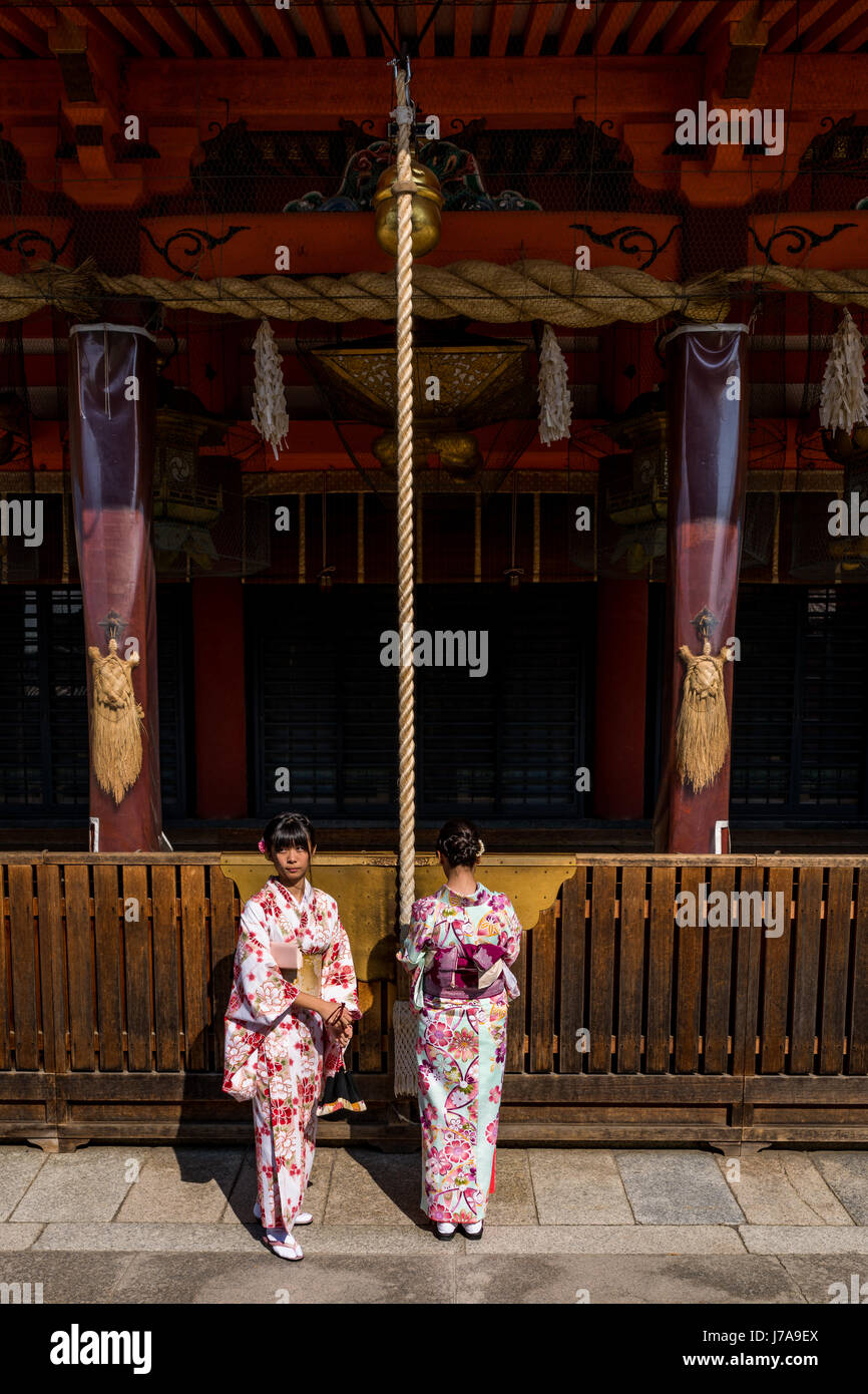 Young Japanese girls wearing furisode, stand at the Yasaka Shrine. One girl is facing the temple while pulling on the bell-rope as a sign of devotion. Stock Photo