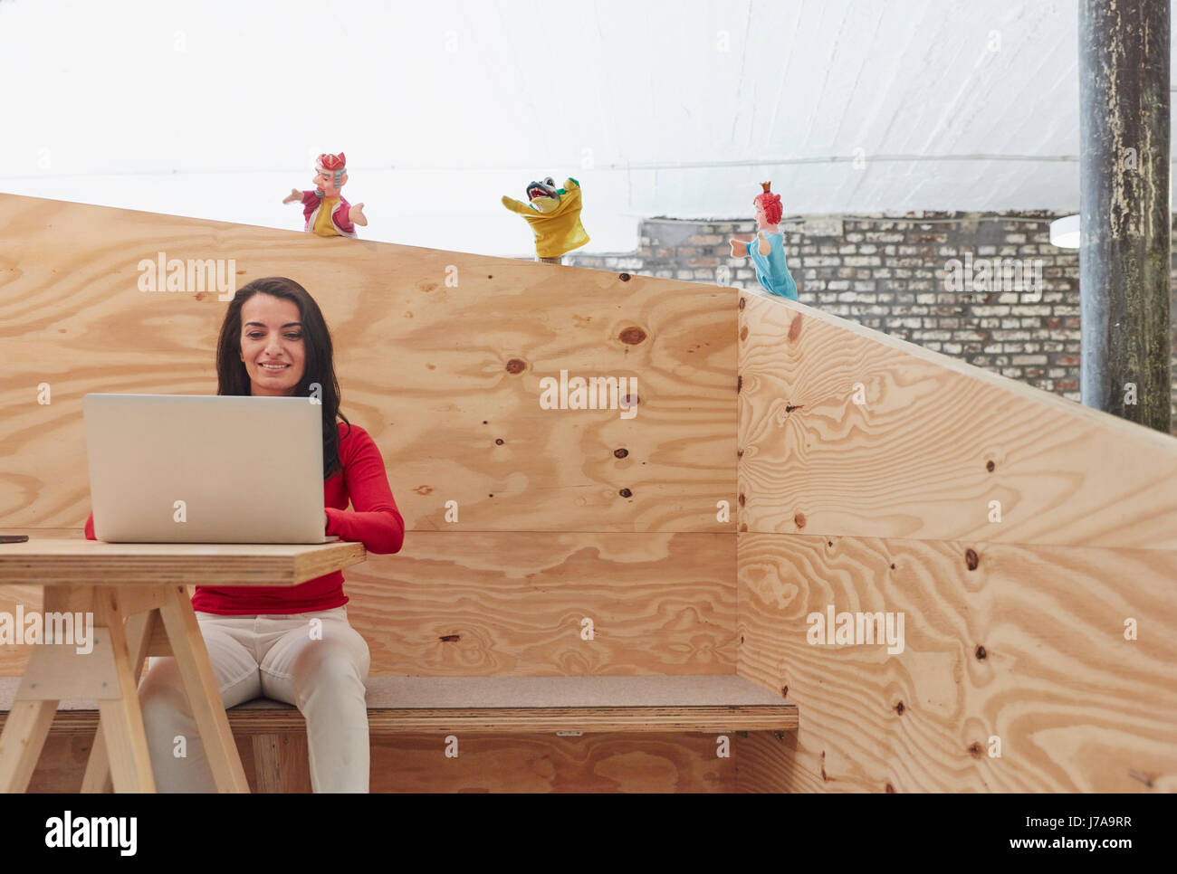 Businesswoman working on laptop with hand puppets watching him from wooden wall Stock Photo