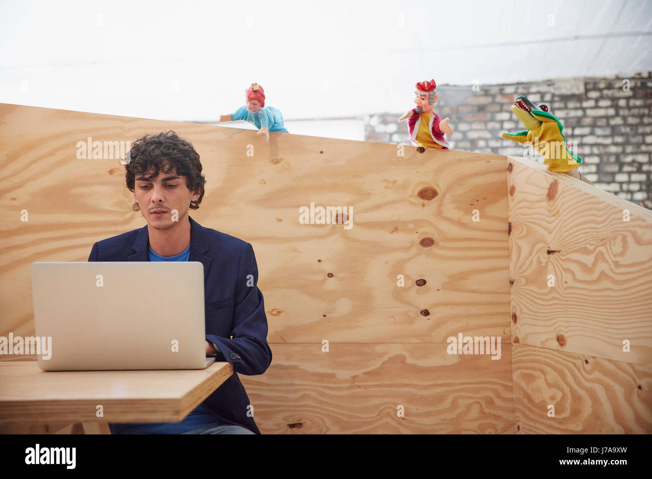 Young businessman working on laptop with hand puppets watching him from wooden wall Stock Photo