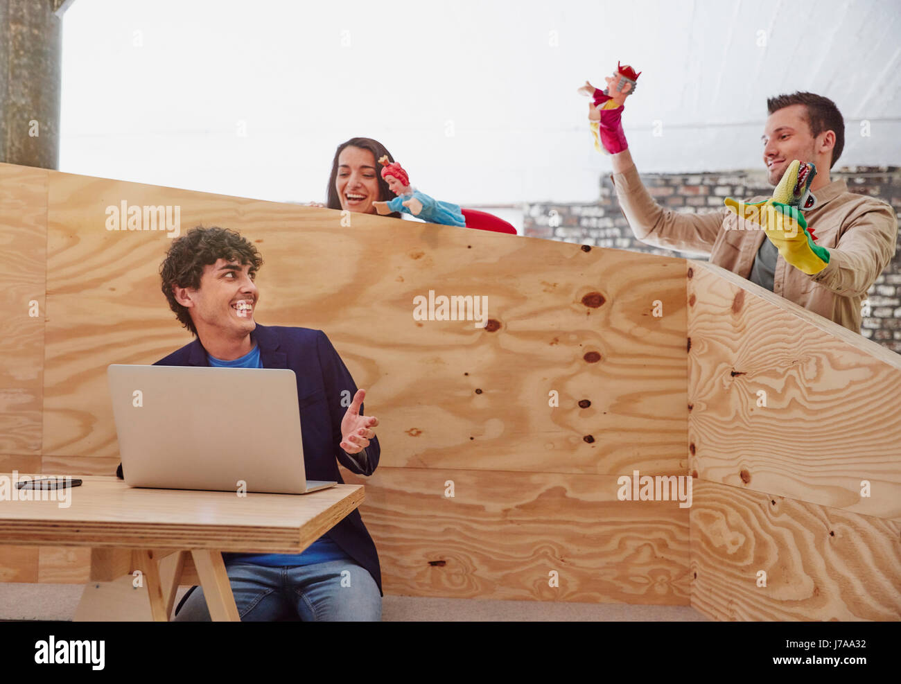 Young businessman working on laptop with colleagues playing hand puppets behind wooden wall Stock Photo
