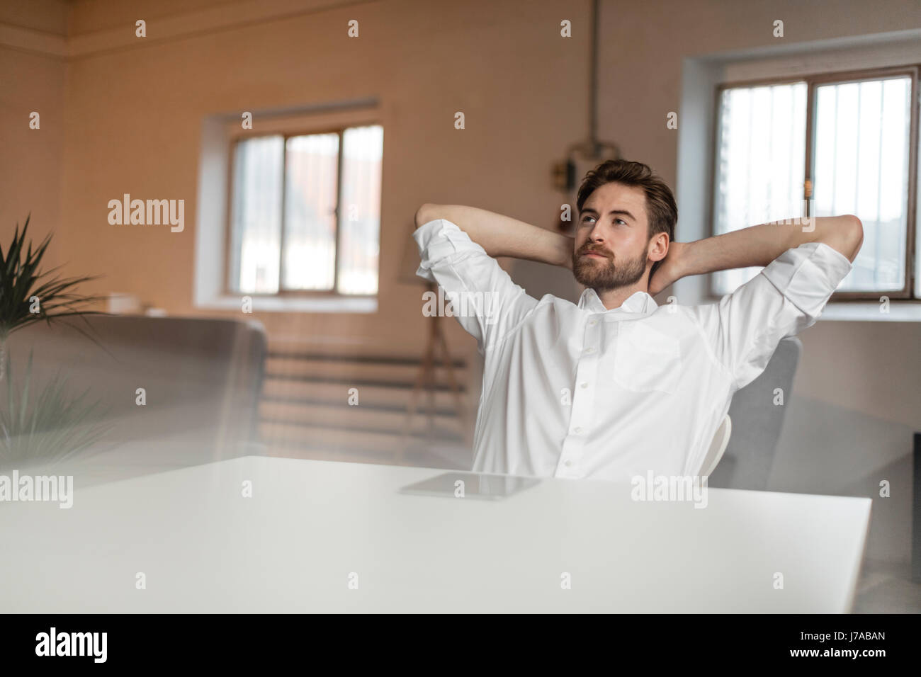 Portrait of pensive young man with hands behind head sitting at table Stock Photo