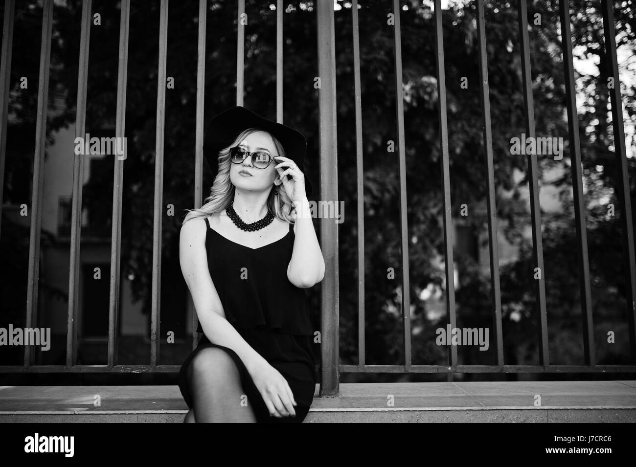 Blonde woman on black dress, sunglasses, necklaces and hat agains iron fence. Stock Photo