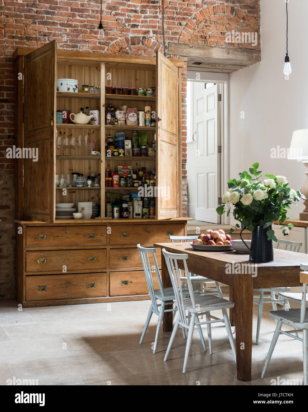 Large pantry cupboard in spacious kitchen with exposed brick wall and farmhouse wooden table Stock Photo