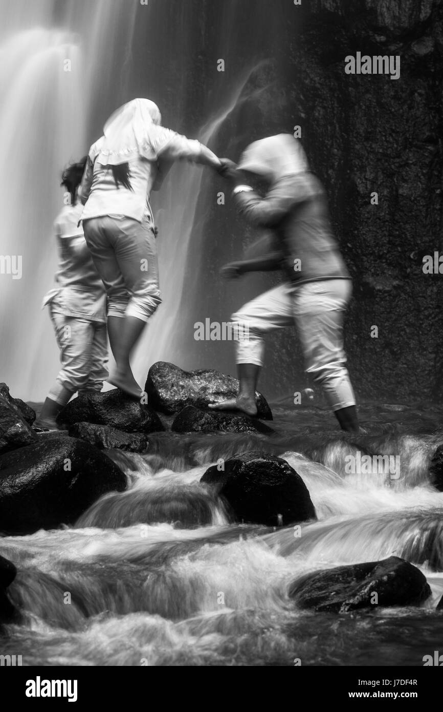 Long exposure black and white photo of Muslim girls helping each other traverse rocky stepping stones at a wild outdoor waterfall in the wilderness Stock Photo