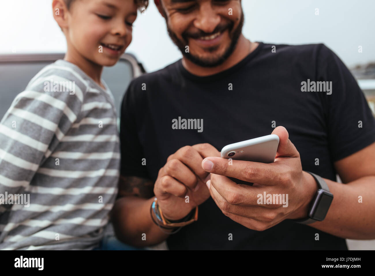Close up of happy father and son in front of the car looking at the mobile phone. Young man and little boy using smart phone while on road trip. Stock Photo