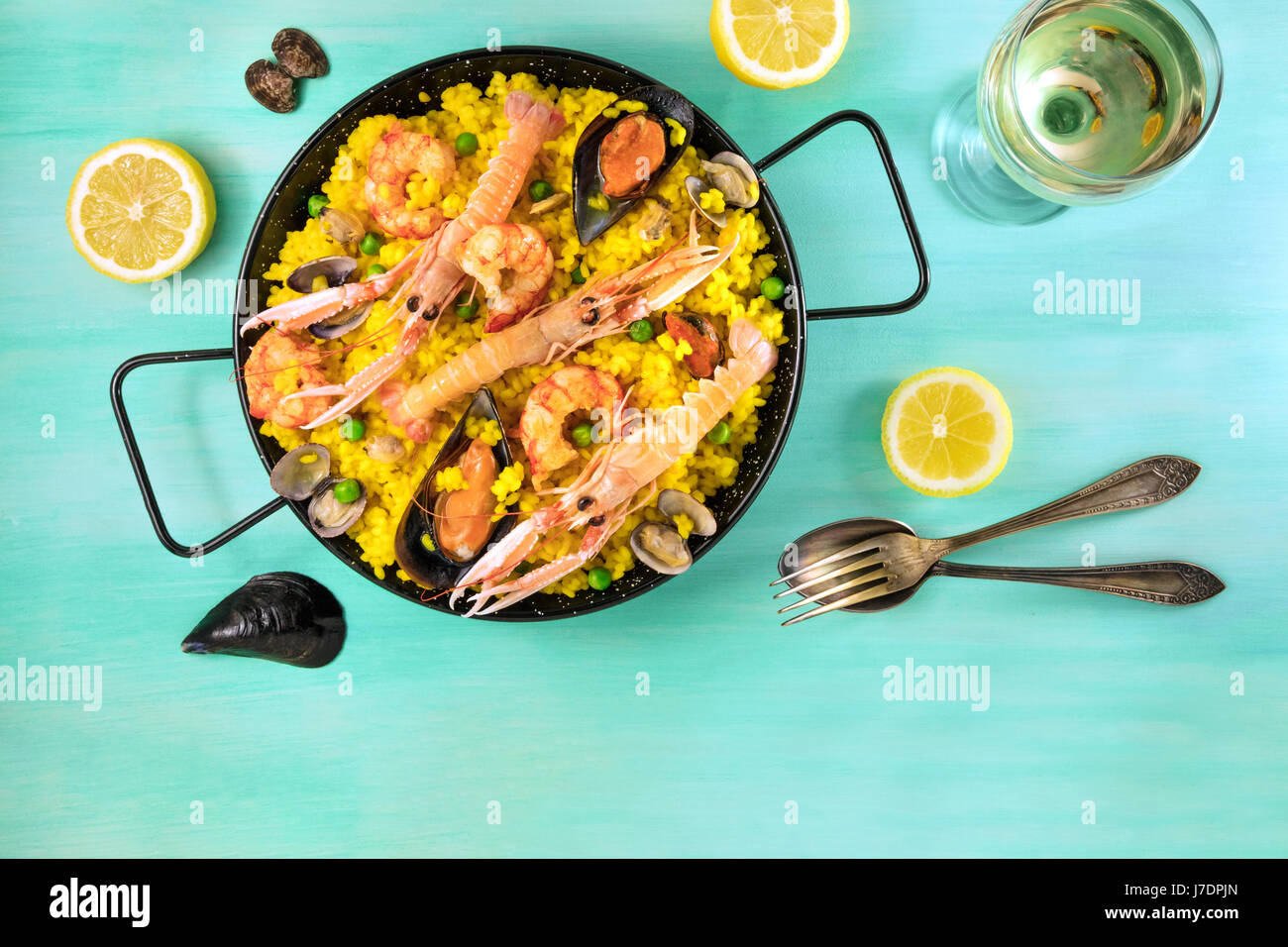 Photo of Spanish seafood paella in typical paellera, with slices of lemons, mussels and clams shells, glass of white wine, fork and spoon, and a place Stock Photo