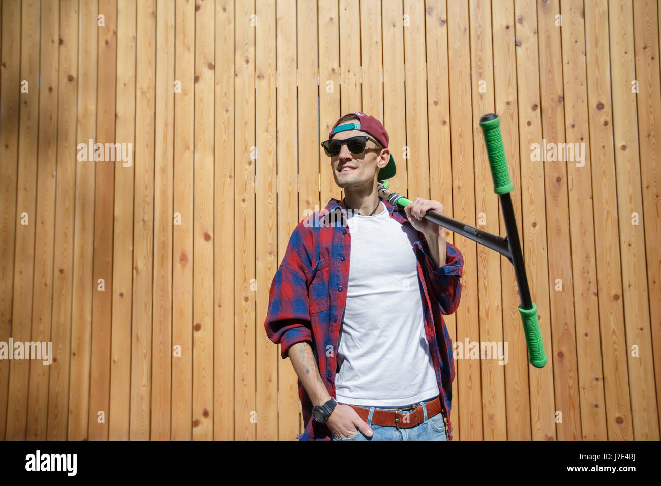 Man looks sideways with scooter on shoulder, standing at wooden fence in spring Stock Photo