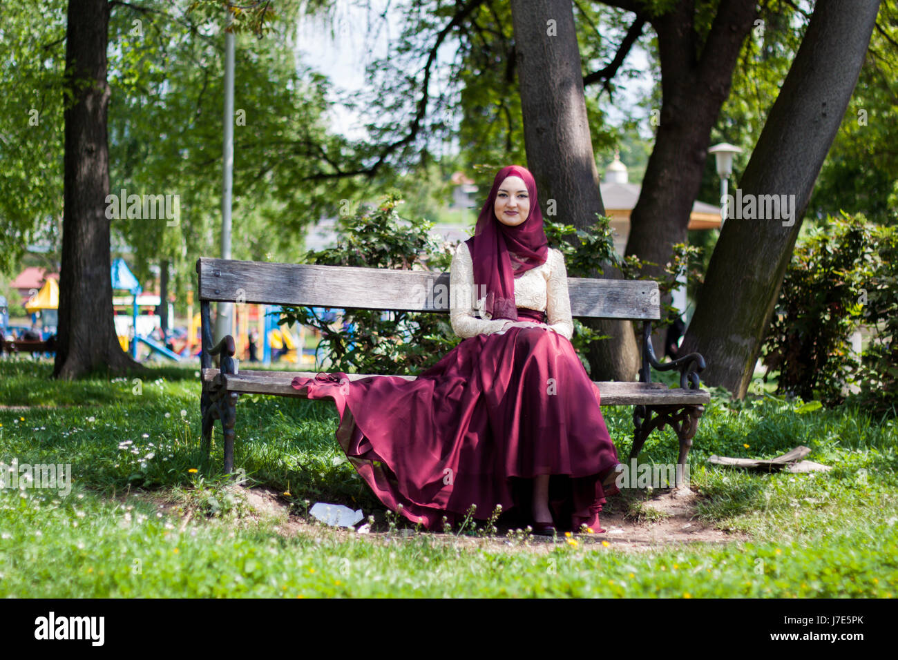 Beautiful girl covered in a beautiful dress sits in a park on a bench ...