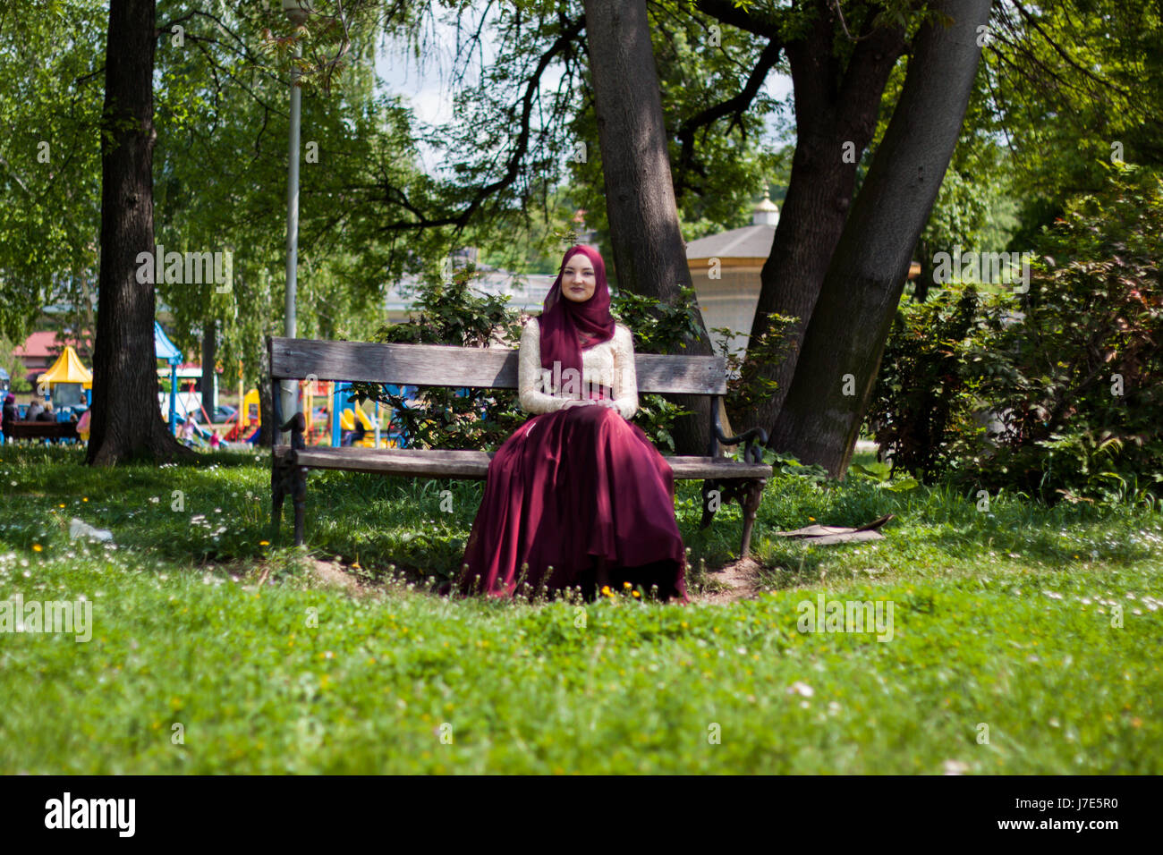 Beautiful girl covered in a beautiful dress sits in a park on a bench ...