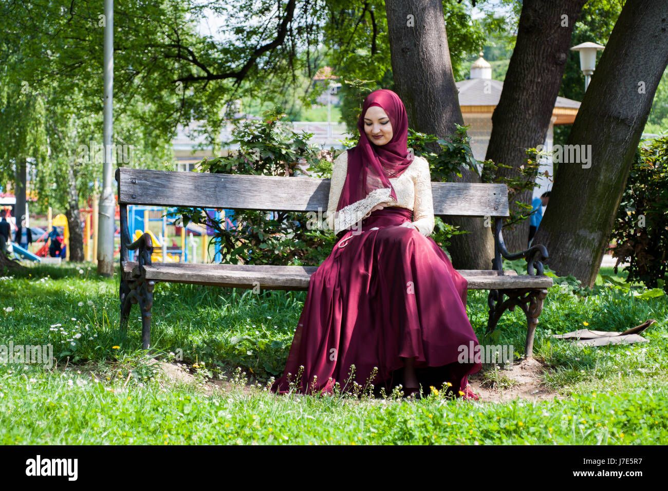 Beautiful girl covered in a beautiful dress sits in a park on a bench ...