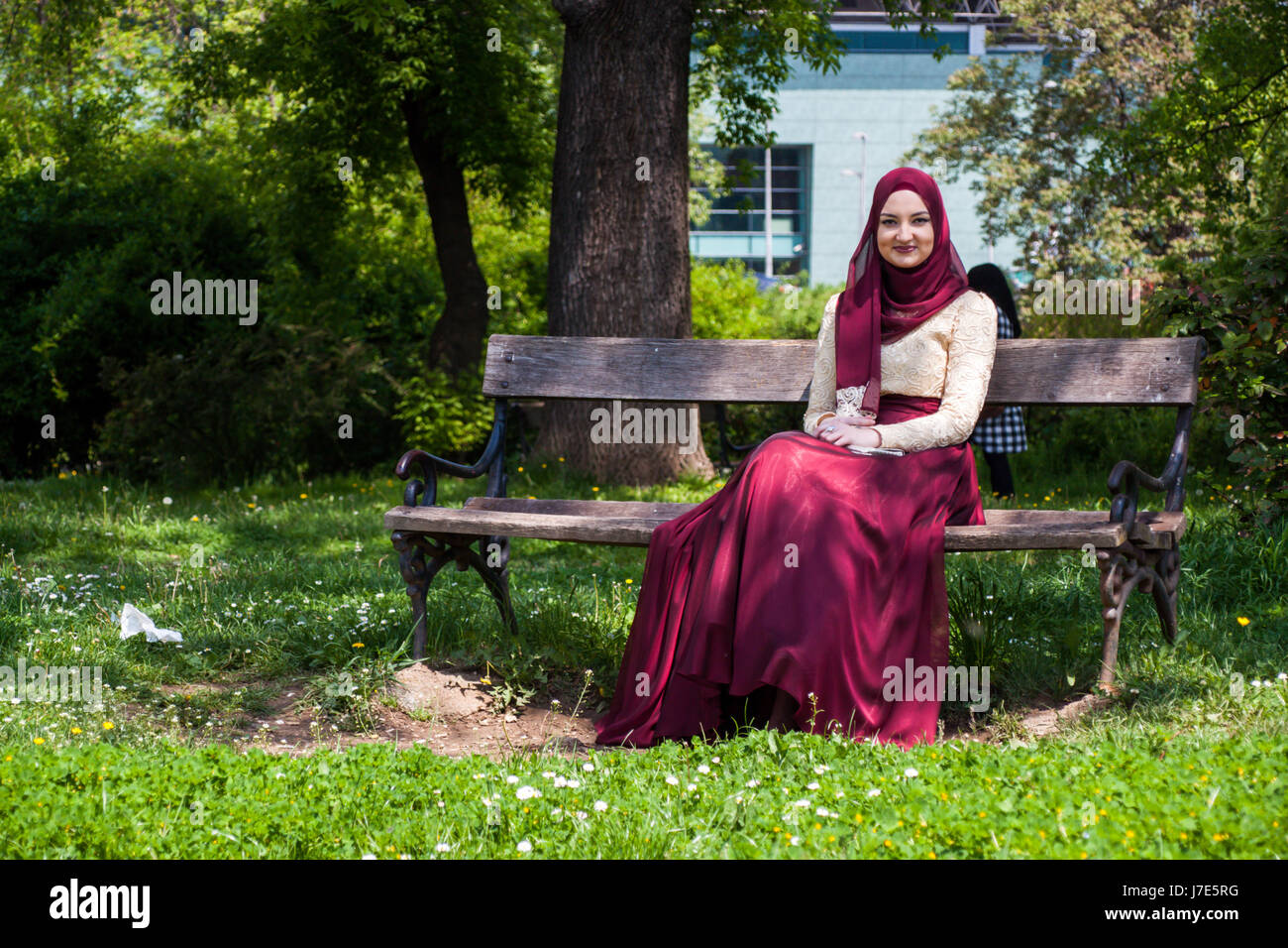 Beautiful girl covered in a beautiful dress sits in a park on a bench ...