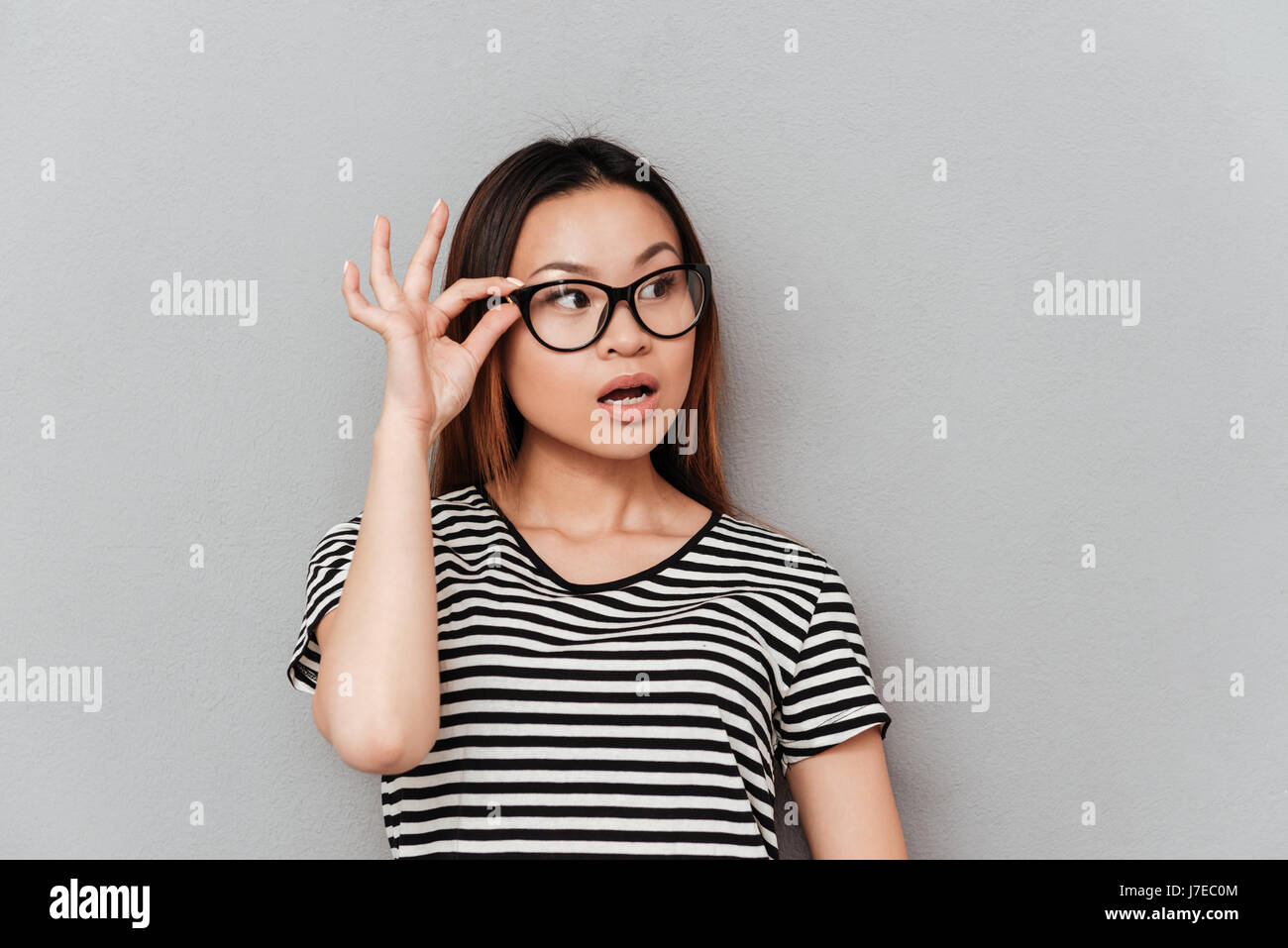 Shocked asian woman looking away and holding her glasses isolated over grey Stock Photo