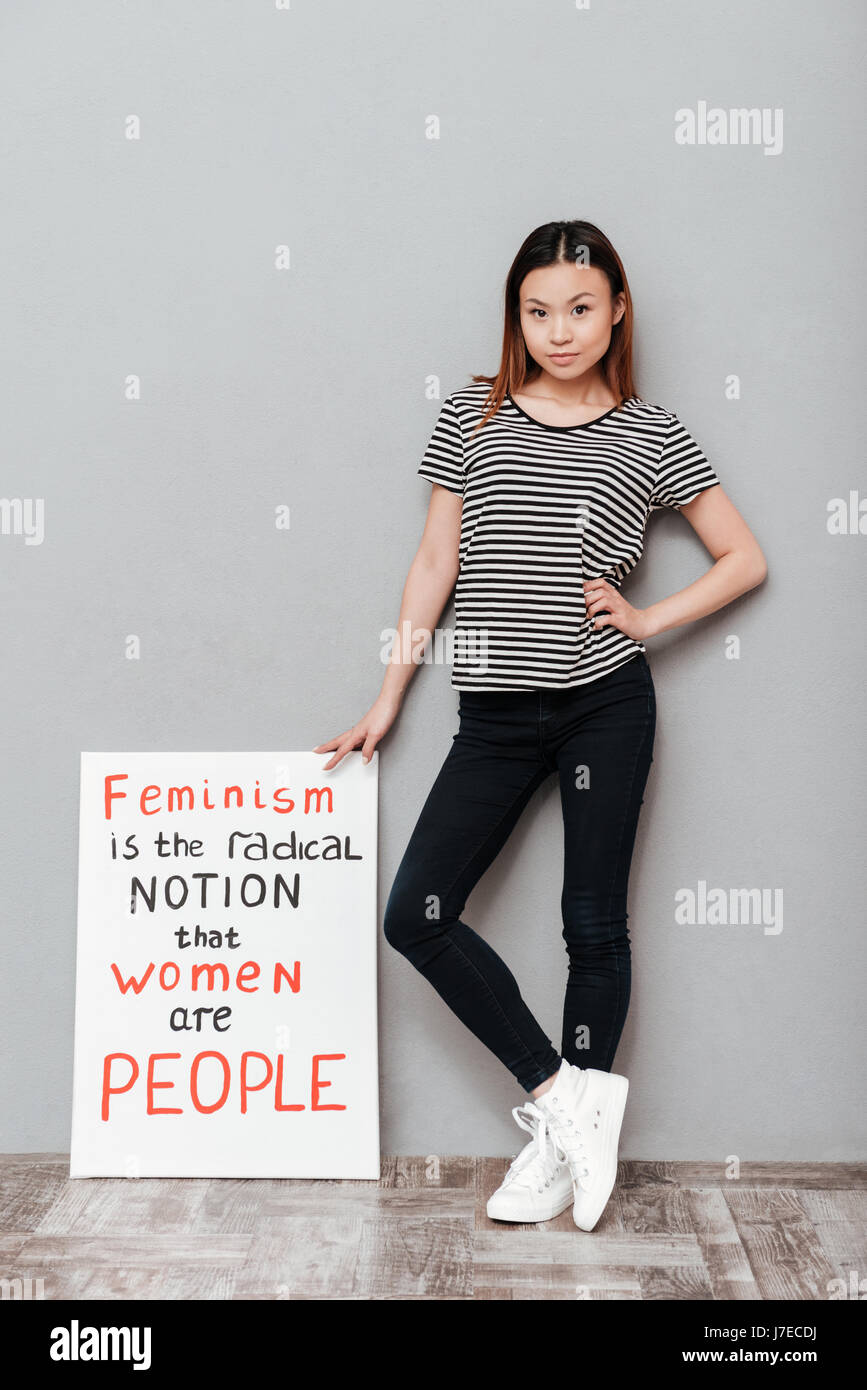 Image of young serious woman standing over grey wall. Looking at camera holding blank with text about feminism. Stock Photo