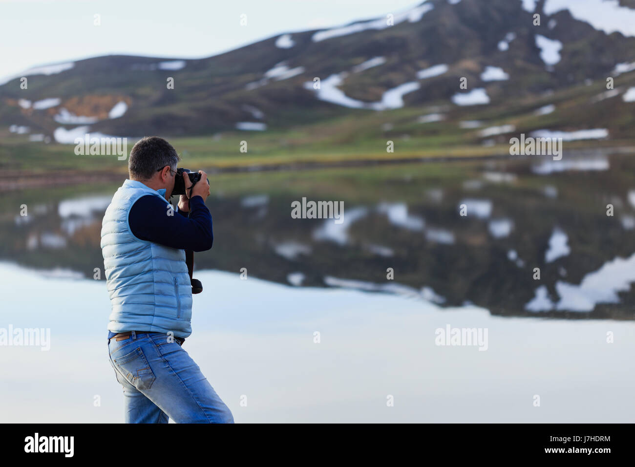 Horizontal sideview portrait of landscape photographer wearing blue jeans and pale blue vest photographing reflections of mountains with melting snow  Stock Photo
