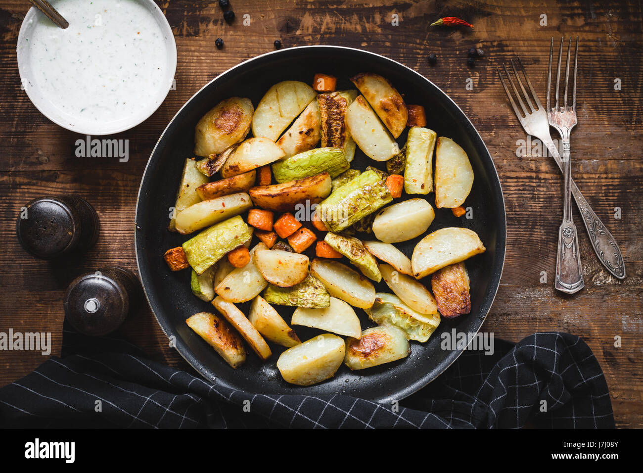 Roasted vegetables, potato wedges, carrots, onion, garlic and zucchini in pan with tzatziki sauce on rustic wooden table. Stock Photo