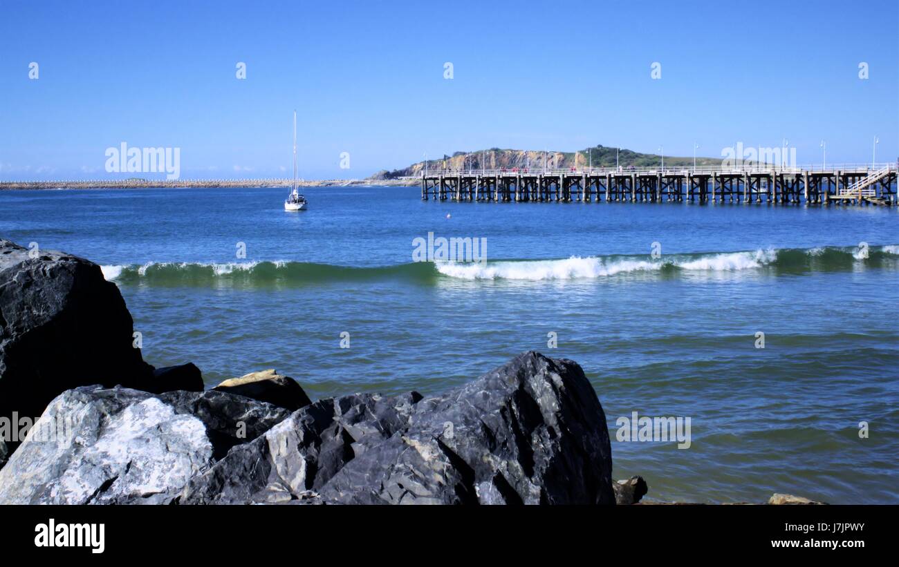 Jetty or Pier over sea with rocks, waves, yacht, boat, hill far in the distance. View of Coffs Harbour Jetty in Australia Stock Photo